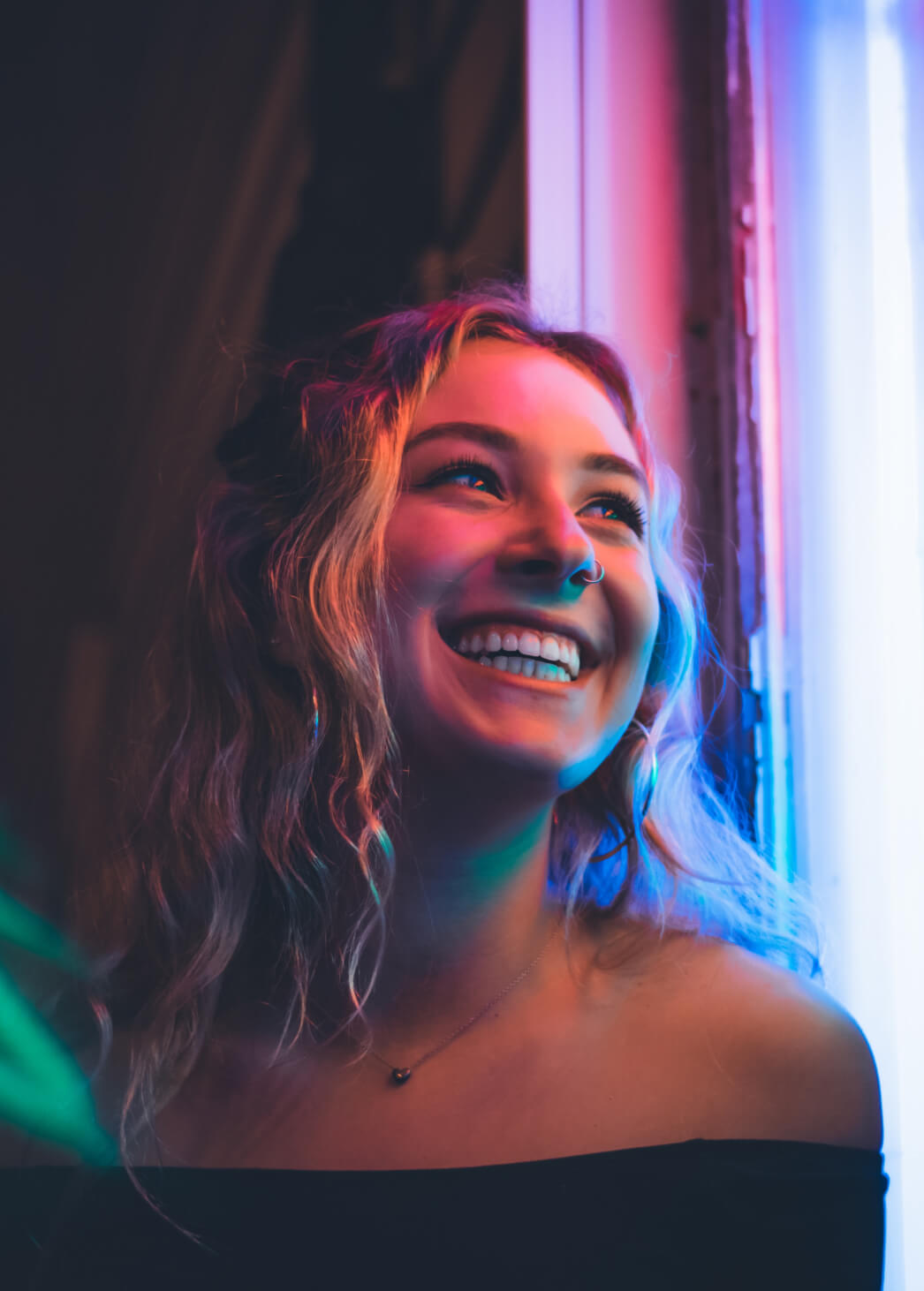 Young blonde woman smiles happily looking out a window with neon lighting bouncing off her