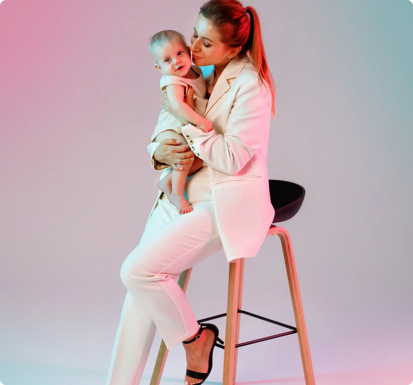 Caucasian woman wearing a white suit with her hair in a ponytail holding her baby sitting on a stool
